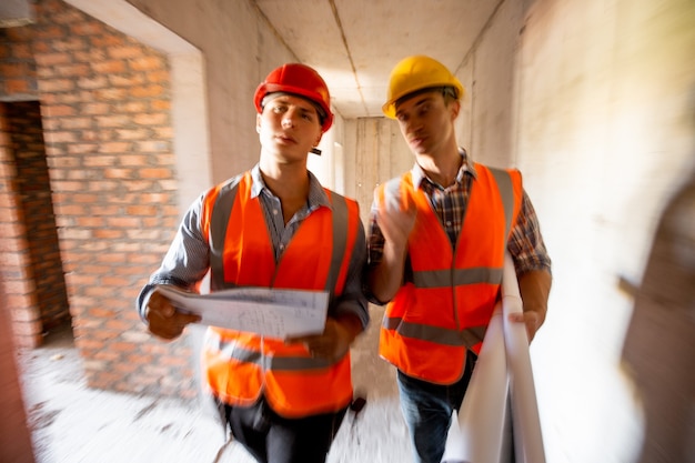 Dos ingenieros civiles vestidos con chalecos de trabajo naranja y cascos caminan dentro del edificio en construcción.