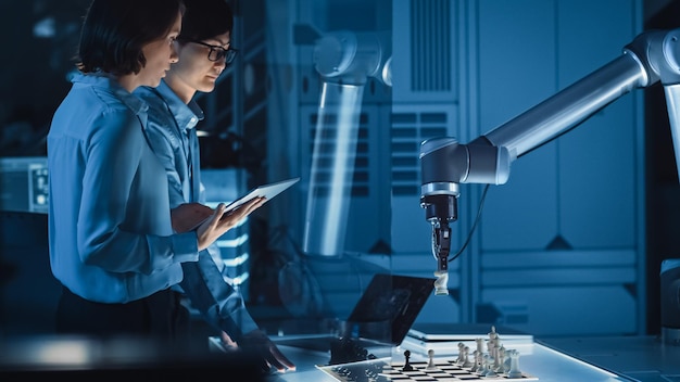 Dos ingenieros de desarrollo están discutiendo y probando una interfaz de inteligencia artificial jugando al ajedrez con un brazo robótico futurista están en un laboratorio de investigación moderno de alta tecnología