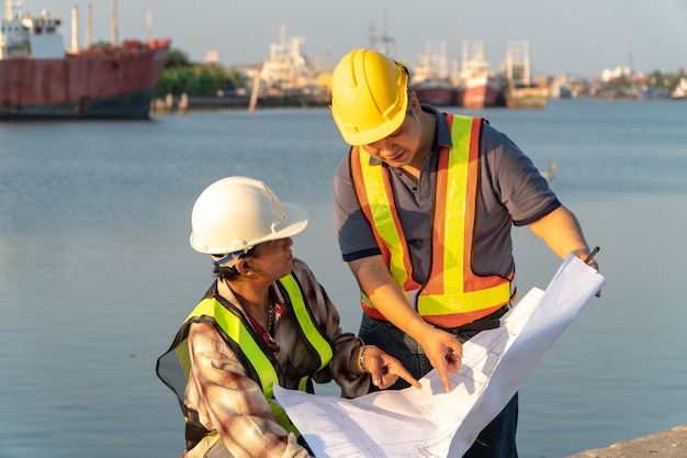 Dos ingenieros estaban de pie con un casco de seguridad. De pie en el muelle Y sosteniendo el plano Y consultando el plano En la construcción. Concepto de gestión eficiente de la obra.