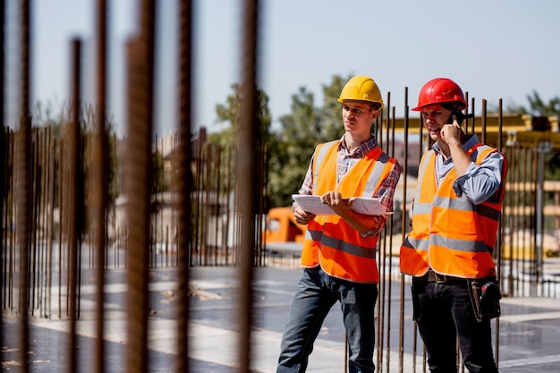 Dos ingenieros estructurales vestidos con camisas, chalecos de trabajo naranjas y cascos exploran la documentación de la construcción y hablan por teléfono en el sitio de construcción cerca de los marcos de acero.