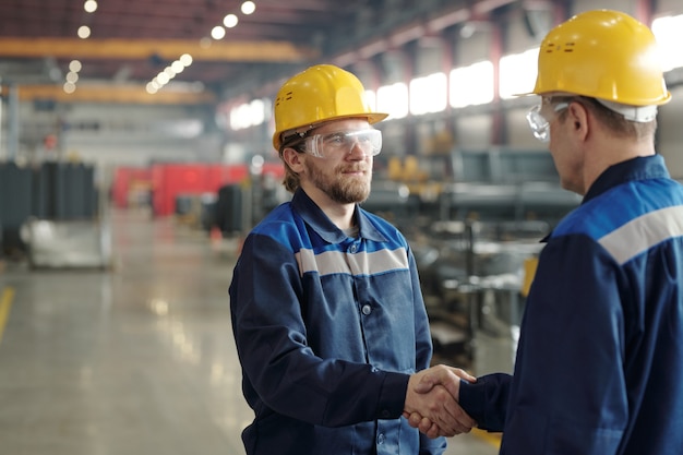 Dos ingenieros masculinos en cascos protectores y ropa de trabajo dándose la mano unos a otros contra el entorno de un gran taller después de hacer un trato