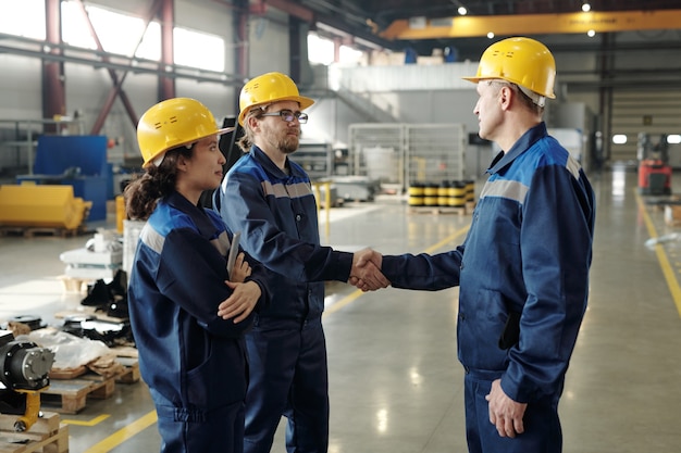 Dos ingenieros masculinos en cascos y ropa de trabajo saludándonos por apretón de manos en el taller con una colega de pie cerca
