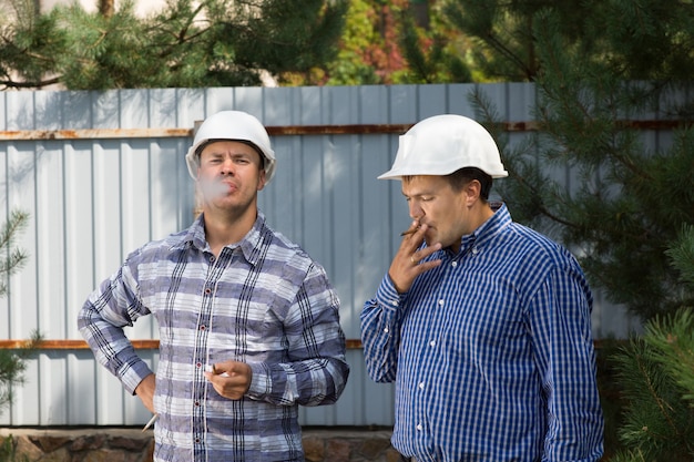 Dos ingenieros tomando un descanso para fumar en sus cascos de pie a la sombra de un árbol en un sitio de construcción fumando sus cigarrillos