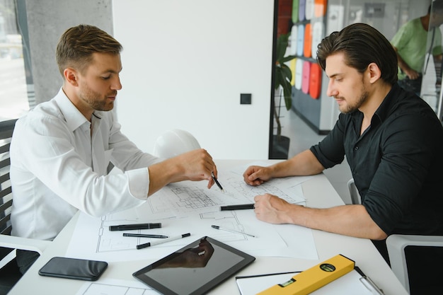 Dos ingenieros trabajando juntos y usando una tableta digital que busca planos y análisis con un plan arquitectónico en el escritorio