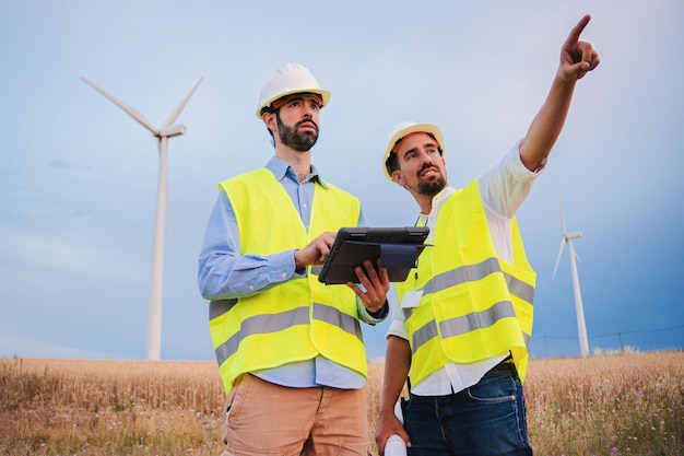 Dos ingenieros de turbinas de molinos de viento trabajando juntos, un técnico que usa un dispositivo de tableta para comprobar la eficiencia, su capataz señala qué generador necesita ser reparado Concepto de energía renovable