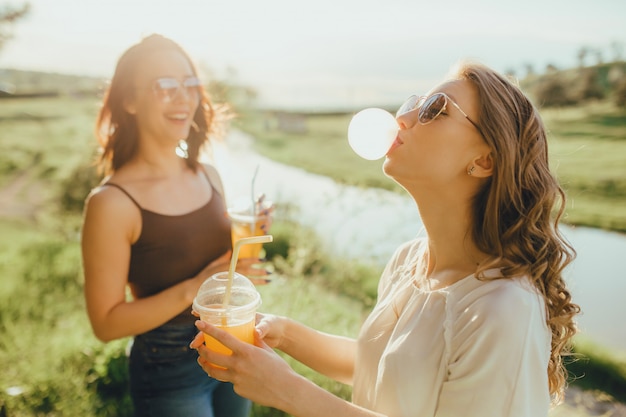 Dos joven explotando una burbuja de un chicle, bebiendo jugo de naranja en un vaso de plástico, al atardecer, expresión facial positiva, al aire libre. infla un chicle.