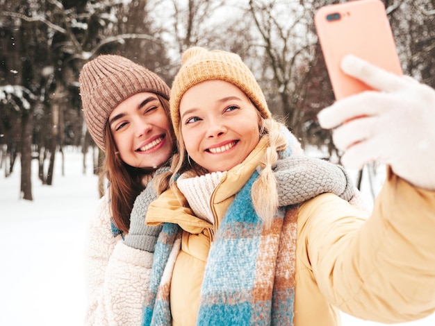 Dos joven hermosa mujer hipster sonriente en ropa de abrigo de moda y bufandas. Mujeres despreocupadas posando en la calle en el parque. Modelos puros positivos divirtiéndose en la nieve. Disfrutando de los momentos de invierno.