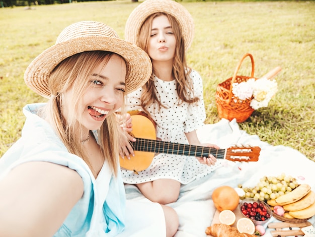Dos joven hermosa mujer hipster en vestidos de verano de moda y sombreros. Mujeres despreocupadas haciendo picnic afuera.