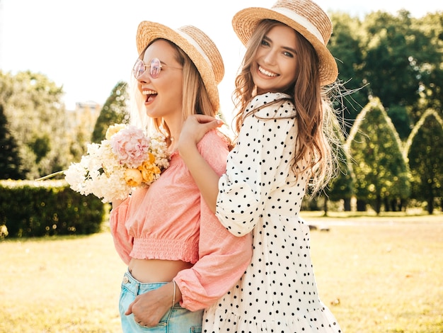 Dos joven hermosa mujer sonriente hipster en vestido de verano de moda. Mujeres despreocupadas sexy posando en el parque con sombreros.