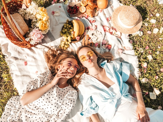 Dos joven hermosa mujer sonriente hipster en vestido de verano de moda y sombreros. Mujeres despreocupadas haciendo picnic afuera.