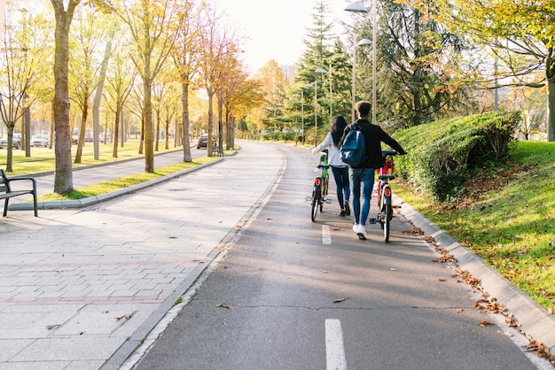 Dos joven pareja caminando por un carril bici con una bicicleta eléctrica para compartir e-bike en sus manos en un hermoso parque con muchos árboles al atardecer