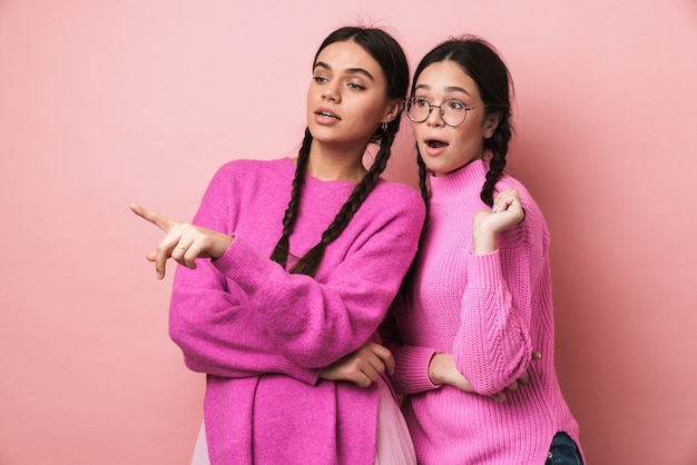 Dos jóvenes adolescentes con trenzas mirando a un lado y señalando con el dedo copyspace aislado sobre pared rosa