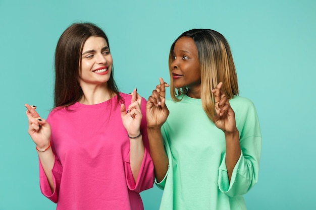 Foto dos jóvenes amigas afroamericanas europeas vestidas de verde rosa manteniendo los dedos cruzados aislados en un retrato de estudio de fondo de pared turquesa azul. concepto de estilo de vida de las personas. simulacros de espacio de copia