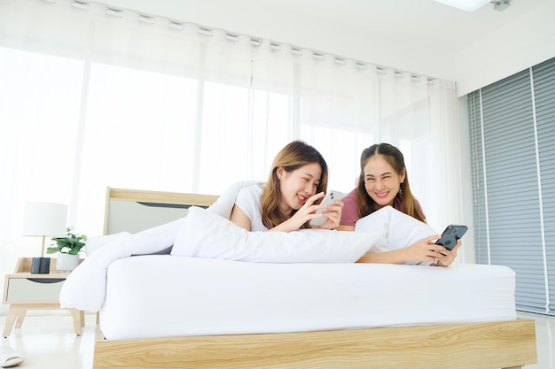 Dos jóvenes amigas asiáticas que usan teléfonos inteligentes mientras se acuestan con una almohada y una manta en la cama juntas Amistad de novia LGBTQIA compartiendo momentos felices juntos en el dormitorio