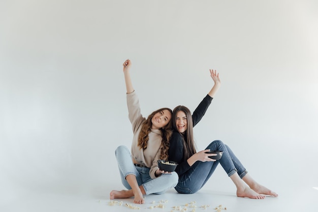 Dos jóvenes amigas bastante alegres que se encuentran aisladas sobre fondo blanco, comiendo palomitas de maíz