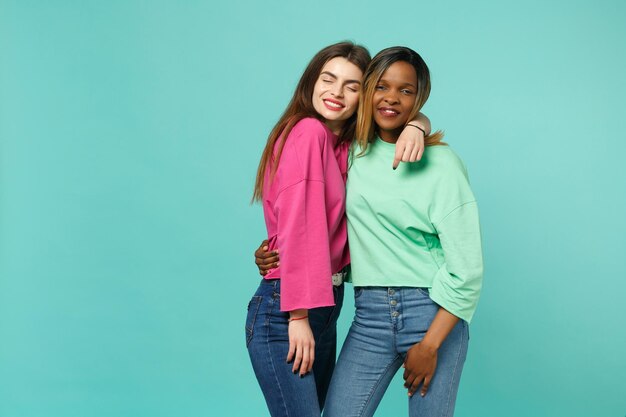 Dos jóvenes amigas europeas y afroamericanas con ropa verde rosa de pie posando aisladas en el fondo azul turquesa de la pared, retrato de estudio. Concepto de estilo de vida de las personas. Simulacros de espacio de copia.