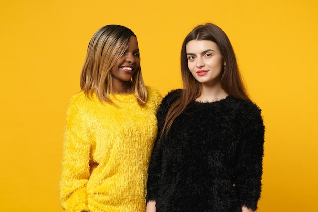 Dos jóvenes amigas europeas y afroamericanas vestidas de amarillo negro de pie posando aisladas en un fondo de pared naranja brillante, retrato de estudio. Concepto de estilo de vida de las personas. Simulacros de espacio de copia.