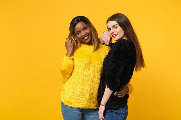 Dos jóvenes amigas europeas y afroamericanas vestidas de amarillo negro de pie posando aisladas en un fondo de pared naranja brillante, retrato de estudio. Concepto de estilo de vida de las personas. Simulacros de espacio de copia.