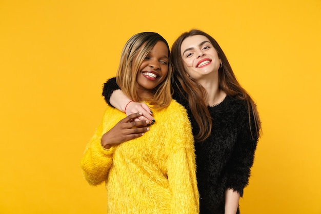 Dos jóvenes amigas europeas y afroamericanas vestidas de amarillo negro de pie posando aisladas en un fondo de pared naranja brillante, retrato de estudio. Concepto de estilo de vida de las personas. Simulacros de espacio de copia.