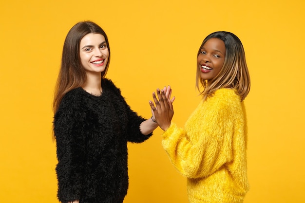 Dos jóvenes amigas europeas y afroamericanas vestidas de amarillo negro de pie posando aisladas en un fondo de pared naranja brillante, retrato de estudio. Concepto de estilo de vida de las personas. Simulacros de espacio de copia.