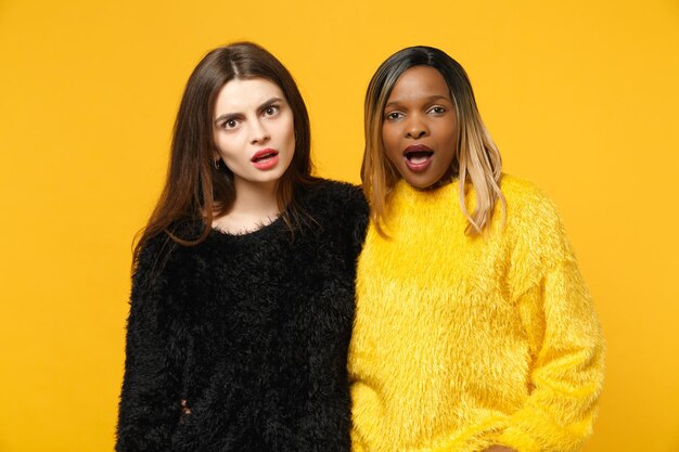 Dos jóvenes amigas europeas y afroamericanas vestidas de amarillo negro de pie posando aisladas en un fondo de pared naranja brillante, retrato de estudio. Concepto de estilo de vida de las personas. Simulacros de espacio de copia.