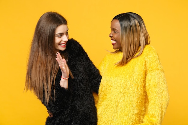 Dos jóvenes amigas europeas y afroamericanas vestidas de amarillo negro de pie posando aisladas en un fondo de pared naranja brillante, retrato de estudio. Concepto de estilo de vida de las personas. Simulacros de espacio de copia.