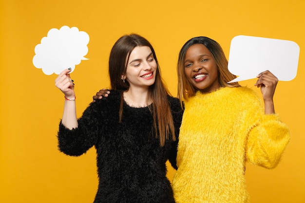 Dos jóvenes amigas europeas y afroamericanas vestidas de amarillo negro sostienen nubes aisladas en un fondo de pared naranja brillante, retrato de estudio. Concepto de estilo de vida de las personas. Simulacros de espacio de copia.