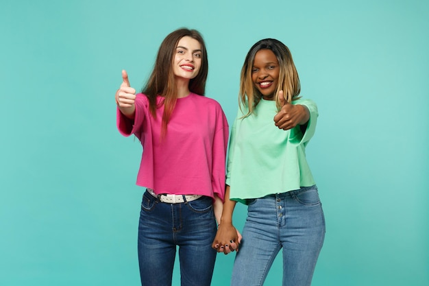 Dos jóvenes amigas europeas y afroamericanas vestidas de verde rosa mostrando los pulgares hacia arriba aisladas en el fondo azul turquesa de la pared, retrato de estudio. Concepto de estilo de vida de las personas. Simulacros de espacio de copia.