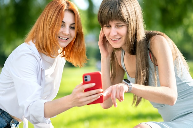 Dos jóvenes amigas sentadas en un banco en el parque de verano tomando un selfie por la cámara del teléfono inteligente.