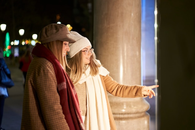 Dos jóvenes amigos mirando en un escaparate, en una ciudad de noche. Uno de ellos señala algo.