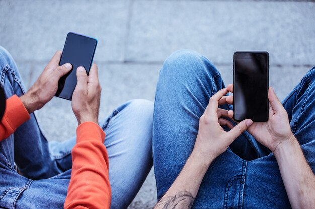 Dos jóvenes amigos en ropa casual con teléfonos inteligentes y computadora portátil en las escaleras al aire libre