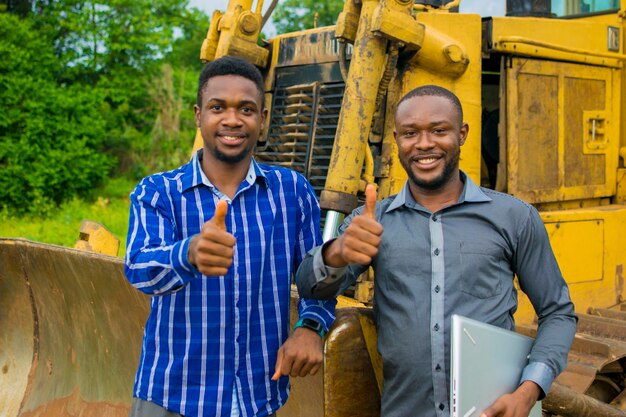 Dos jóvenes apuestos contratistas africanos se sienten abrumados mientras están parados junto a un tractor e hicieron los pulgares hacia arriba