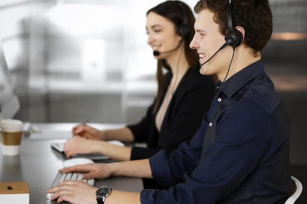 Dos jóvenes con auriculares están hablando con los clientes, mientras están sentados en el escritorio de una oficina moderna.