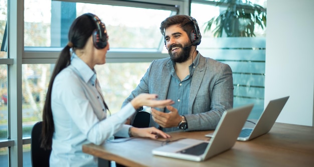 Dos jóvenes con auriculares hablando entre sí mientras están sentados en el escritorio de una oficina Operadores del centro de llamadas en el trabajo