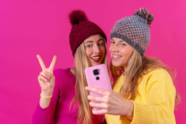 Dos jóvenes caucásicas rubias con un teléfono aislado en un selfie de fondo rosa haciendo el gesto de ok