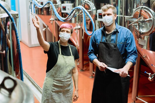 Dos jóvenes cerveceros con máscaras protectoras discutiendo la calidad de los nuevos equipos de producción de cerveza mientras uno de ellos apunta al gran tanque de acero