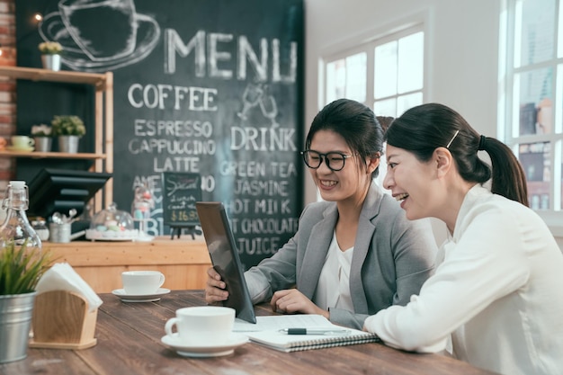 Dos jóvenes colegas asiáticas en la cafetería. empresarias en traje usando tableta con documentos trabajando en la mañana en la cafetería. damas de oficina compañero de trabajo tomando un descanso relajante hablando sonriendo.