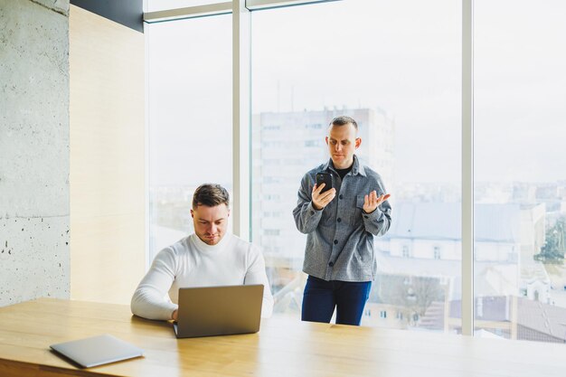 Dos jóvenes confiados están sentados juntos en una mesa y uno de ellos apunta a una computadora portátil Trabajar en un nuevo proyecto Colegas exitosos trabajando juntos en la oficina