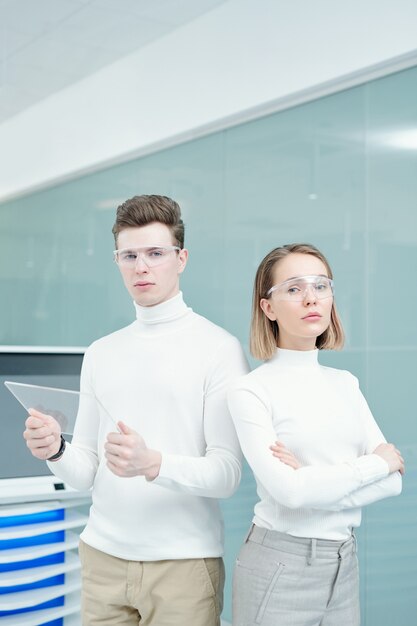 Dos jóvenes consultores del centro de tecnologías modernas serios en puentes blancos y gafas inteligentes de pie delante de la cámara en la oficina