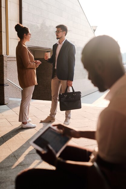 Foto dos jóvenes corredores confiados en ropa casual elegante discutiendo puntos de trabajo en una reunión al aire libre mientras su colega usa el panel táctil