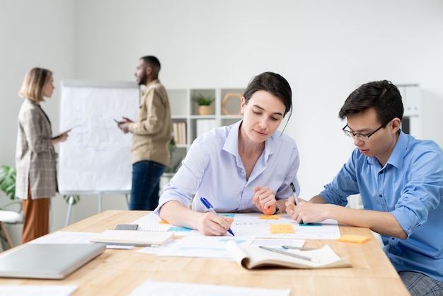 Dos jóvenes corredores interculturales sentados a la mesa en la oficina y analizando datos o haciendo notas de trabajo