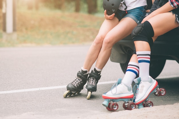 Dos jóvenes delgadas y sexys y patines. Una hembra tiene patines en línea y la otra tiene patines cuádruples. Las chicas cabalgan bajo los rayos del sol.