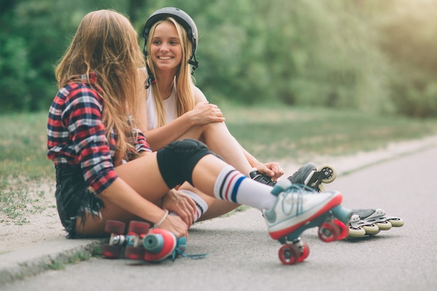 Dos jóvenes delgadas y sexys y patines. Una hembra tiene patines en línea y la otra tiene patines cuádruples. Las chicas cabalgan bajo los rayos del sol.