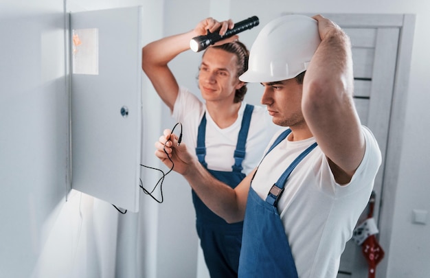 Dos jóvenes electricistas trabajan juntos en interiores usando una linterna