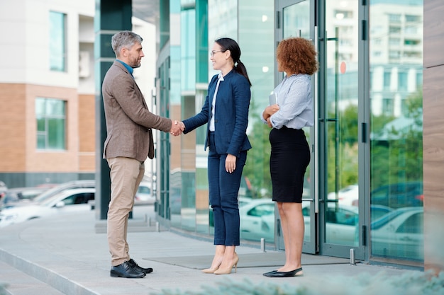 Dos jóvenes empresarias elegantes en ropa formal que se encuentran con su socio comercial extranjero por la entrada del edificio de oficinas