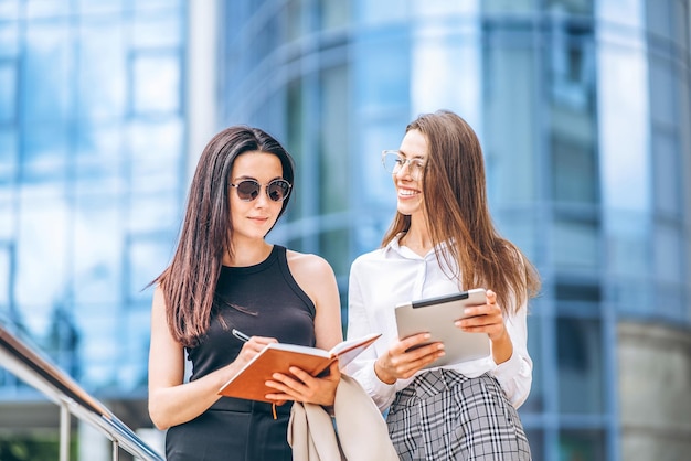 Dos jóvenes empresarias con tableta y portátil caminando al aire libre cerca del moderno centro de negocios