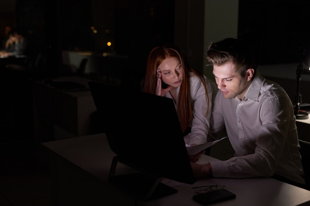 Dos jóvenes empresarios caucásicos trabajando juntos en la computadora, discutiendo el proyecto en la oficina por la noche. vista lateral de la dama pelirroja y los colegas de chico guapo brainsotrming. en la sala de juntas oscura