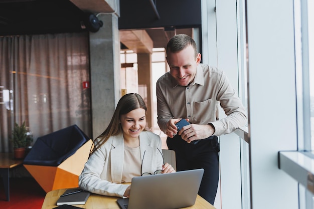 Dos jóvenes empresarios y empresarias sonriendo mientras están sentados en la oficina hablando y navegando por Internet junto con una computadora portátil