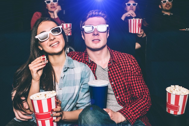 Dos jóvenes están sentados uno cerca del otro. Tienen anteojos especiales en la cara para ver películas. La niña sostiene una canasta con palomitas de maíz y una pieza en sus manos. El hombre tiene una taza de coca