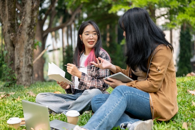 Dos jóvenes estudiantes asiáticas están sentadas en la hierba en un parque estudiando juntas.